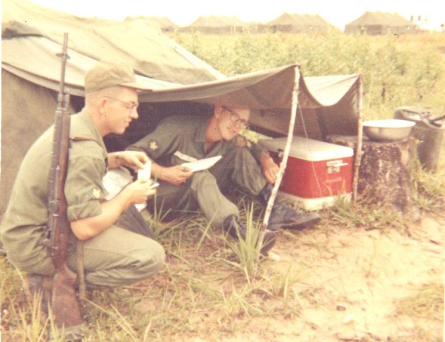 Ronnie, shown on the left, squats near a pup tent made by connecting the ponchos, stakes, and poles of two men. Because of the cooler shown, this might have been taken during training.