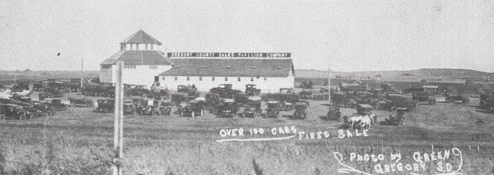 One copy of this photo was dated 1921. The handwritten caption says, “Over 100 cars, first sale.” The sign on the roof identifies the building as Gregory County Sales Pavilion Company.